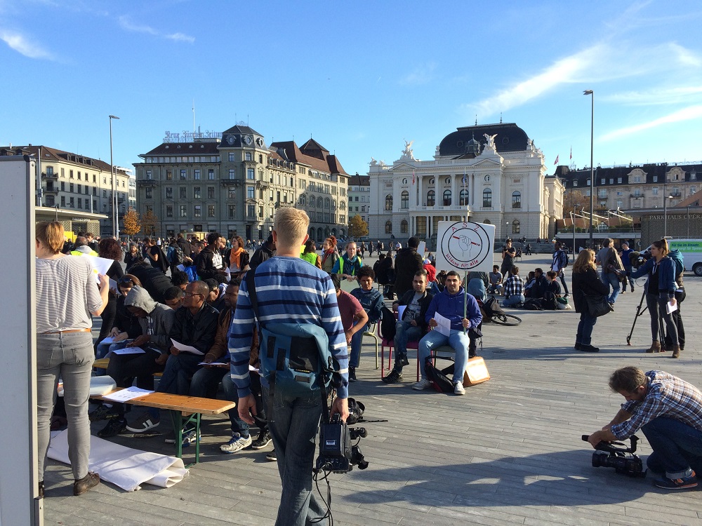 Ein grosser Stadtplatz voller Menschen. In der Nähe sitzt eine Gruppe von Menschen auf Bänken und hört zu, wie eine Person vor einem Whiteboard etwas erklärt.