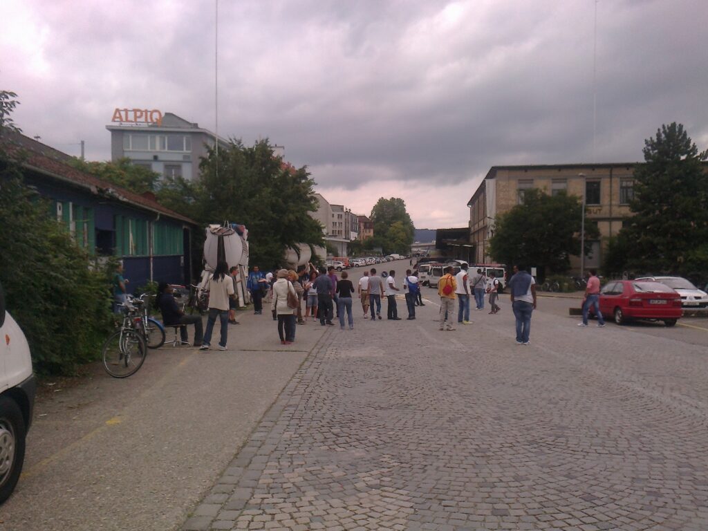 
Eine Gruppe von Menschen steht auf einer breiten Strasse vor dem ASZ-Gebäude. Einige Menschen sprechen miteinander, andere gehen.
