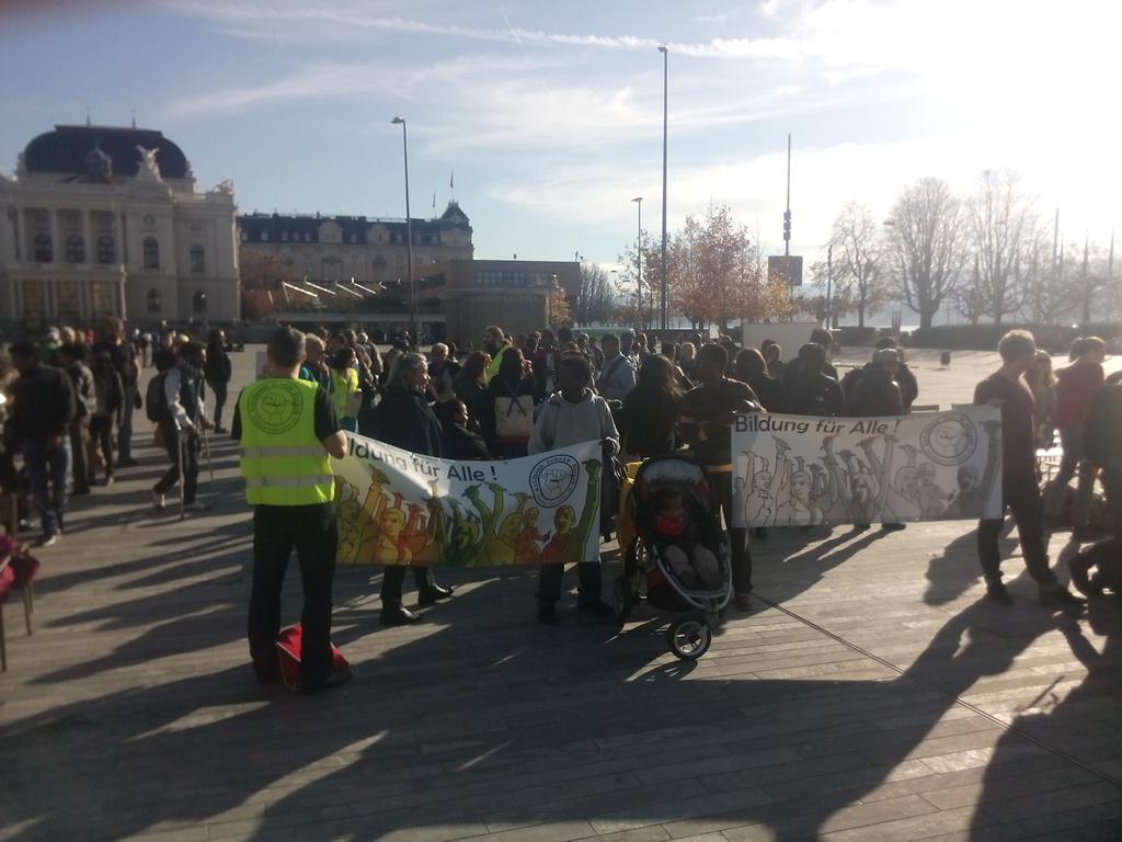  Auf einem sehr großen öffentlichen Platz beobachtet eine Gruppe von Menschen ein Whiteboard. Vor ihnen halten 4 Personen Transparente mit der Aufschrift „Bildung für alle“ in der Hand, und es gibt sogar ein Baby in einem Kinderwagen. 