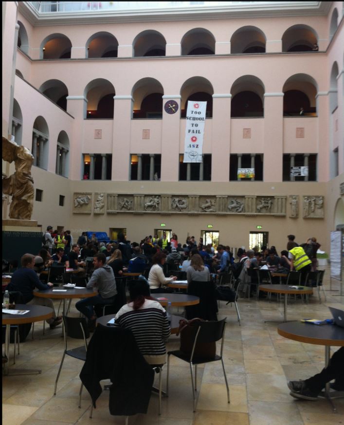 
Many people are sitting around many small roundtables, each one has pen and paper and pays attention to the lesson. They are all in the university atrium. a very large open space with roman-style wall reliefs around them.
