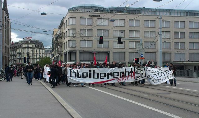 Eine grosse Gruppe von Menschen marschiert die Strasse entlang und trägt ein Transparent mit der Aufschrift „Bleiberecht“.